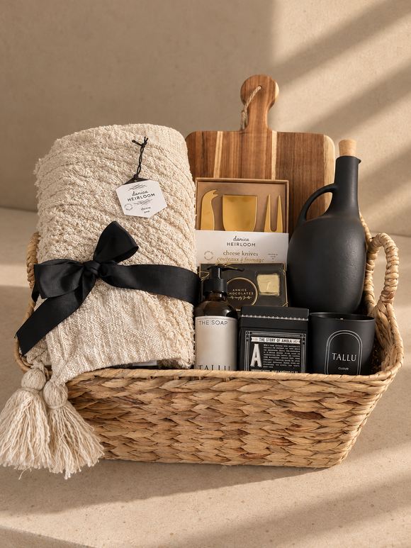 A Warm Welcome housewarming gift basket featuring a woven throw blanket, wooden serving board and cheese knives, olive oil, scented soap, candle, and gourmet chocolates, arranged in a natural woven basket.