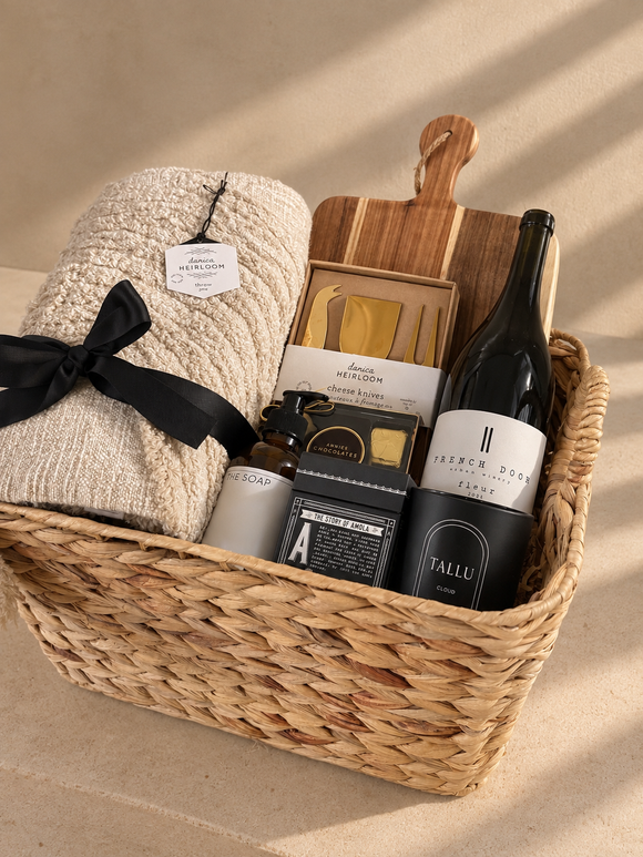A Warm Welcome housewarming gift basket featuring a woven throw blanket, wooden serving board and cheese knives, olive oil, scented soap, candle, and gourmet chocolates, arranged in a natural woven basket.