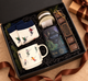 Holiday gift box featuring an All Is Calm tea, festive ski-patterned socks and mug, artisan chocolates, and a scented candle, styled with evergreen accents and brown ribbon.