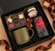 Holiday corporate gift box featuring Christmas tea, a ribbed ceramic mug, artisan chocolates, and a candle, styled with evergreen accents and brown ribbon in a black gift box.