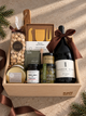 A gift basket from Salt Gift Co. filled with gourmet items, including a bottle of French Door Vin Rouge red wine, almonds, local soy candle, honey, olives popcorn, cheese knife set, arranged in a light wood tray with a brown ribbon.