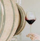 A hand holding a clear wine glass with red wine, set against a blurred background of wooden wine barrels.