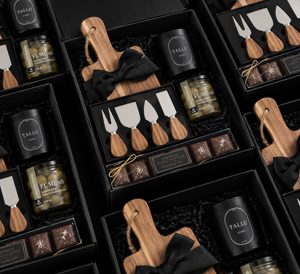 Luxury corporate gift box featuring an acacia serving board, wooden-handle cheese knife set, gourmet olives, and artisan chocolates, arranged in a black gift box with black ribbon.