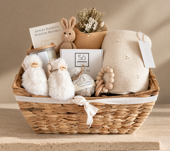 A woven natural-fiber gift basket filled with baby nursery items, including a cream knit bonnet, two white plush duck toys, a small crochet bunny, a jar candle, a box of tea, dried flowers, and a wooden bead teething ring, set against a warm beige background.